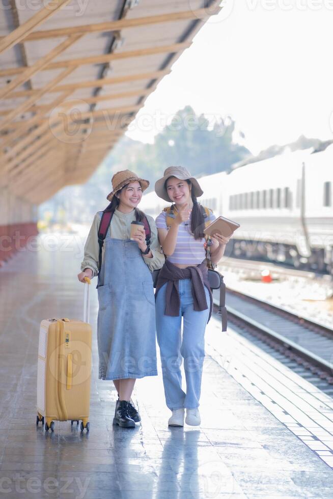 dos joven asiático amigos muchachas con mochilas a ferrocarril estación esperando para tren, dos hermosa mujer caminando a lo largo plataforma a tren estación foto