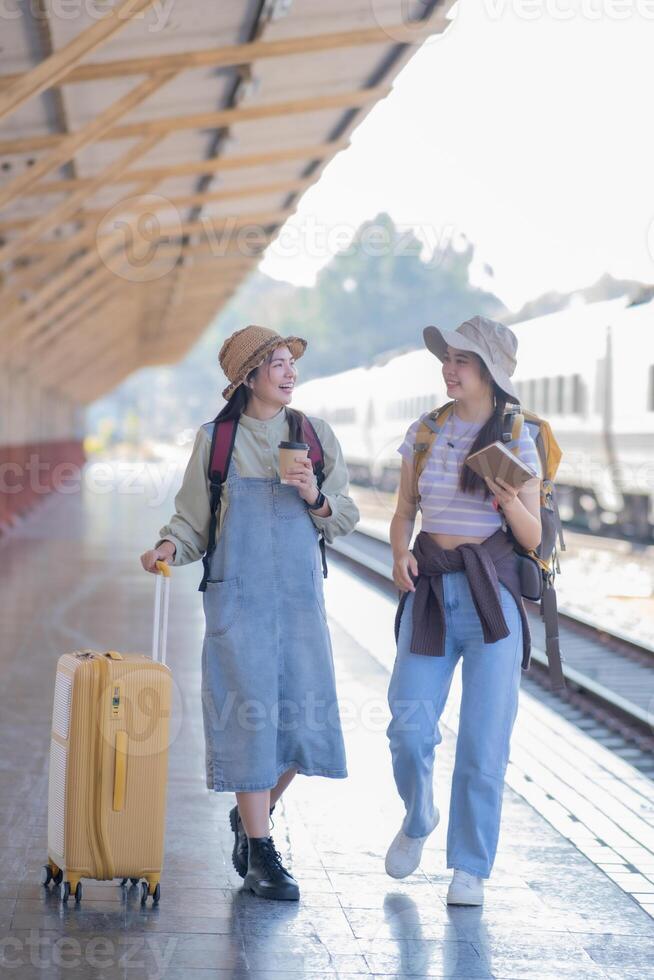 dos joven asiático amigos muchachas con mochilas a ferrocarril estación esperando para tren, dos hermosa mujer caminando a lo largo plataforma a tren estación foto