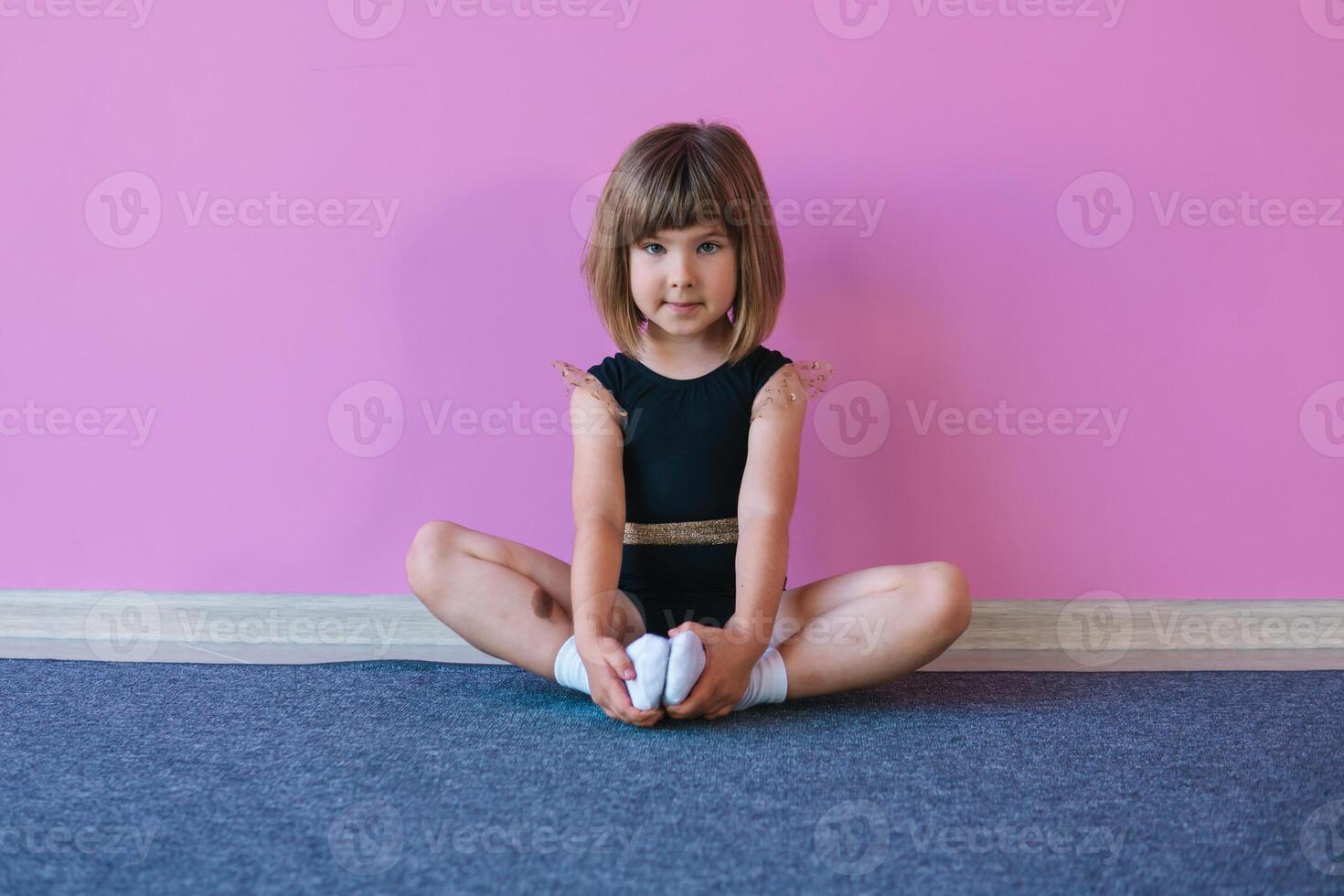 Little gymnast girl with blond hair in a beautiful black leotard is sitting on a background of a pink wall photo