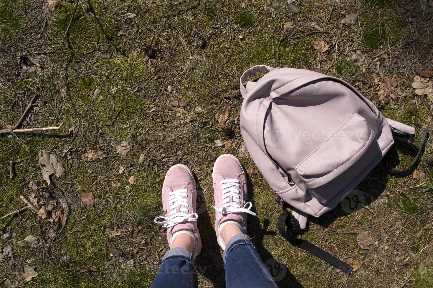 Female legs in jeans and pink sneakers are standing in a ground of forest with backpack. photo