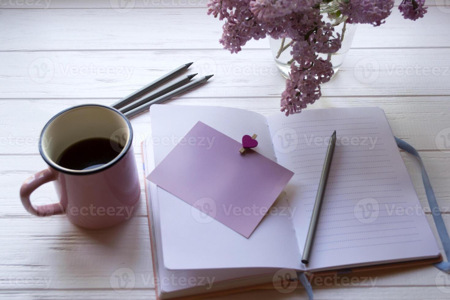 Notepad, pencils, lilac in vase and a cup of coffee on a white wooden desktop. Inspirational workplace. photo