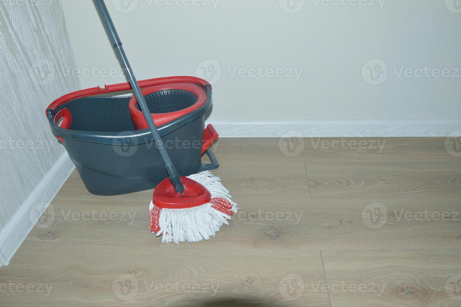 A black bucket with a wringer and a mop stand on a wooden floor near a white wall. Accessories for cleaning home and office. photo