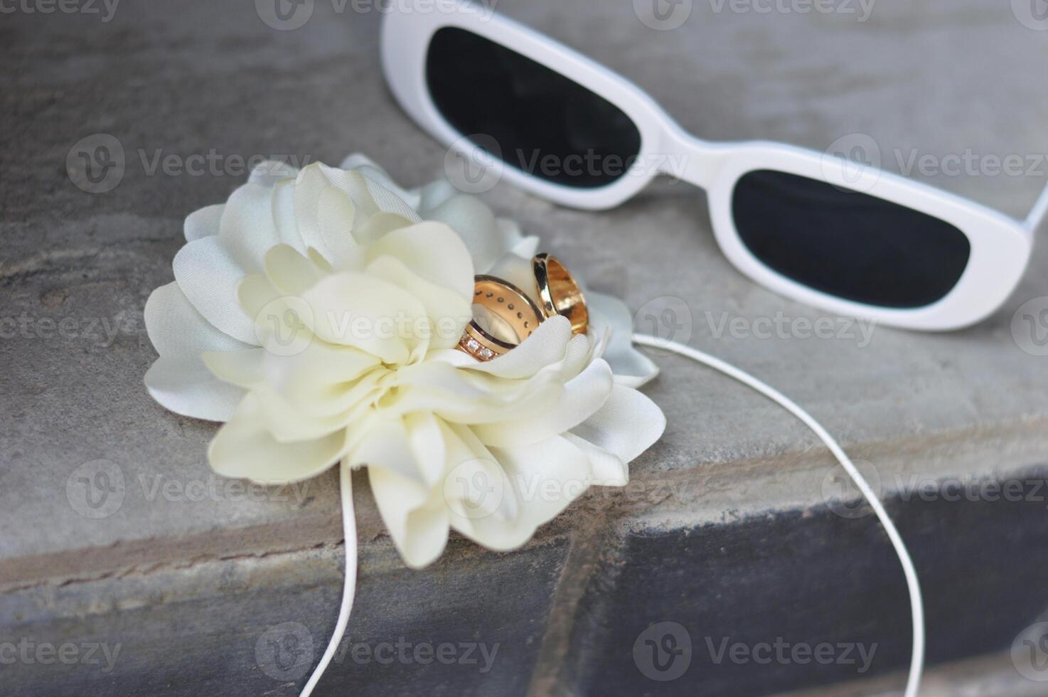 Sunglasses, white boutanier flower and newlyweds rings on a gray concrete surface. Wedding accessories. photo
