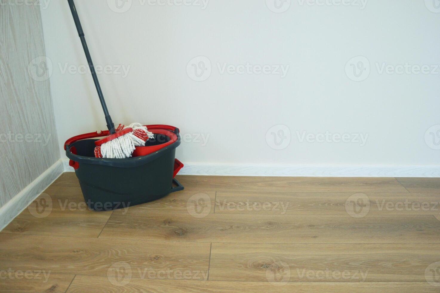 Cleaning mop and black bucket with wringer on wooden floor near white wall. Accessories for cleaning home and office. photo