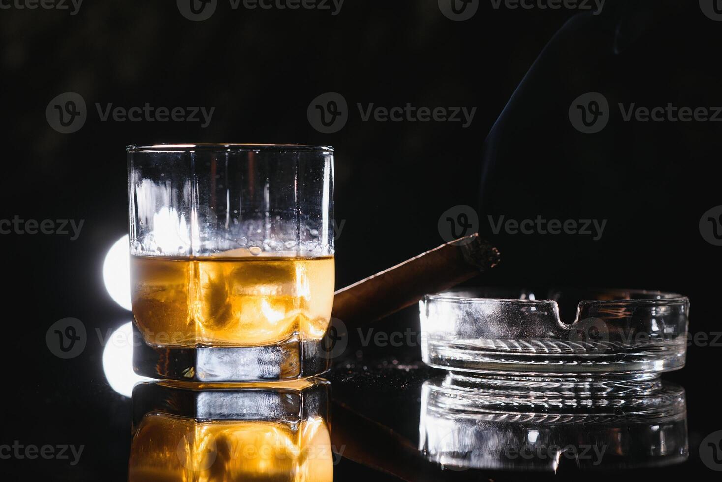 Glass of whiskey with ice cubes and smoking cigar on table photo