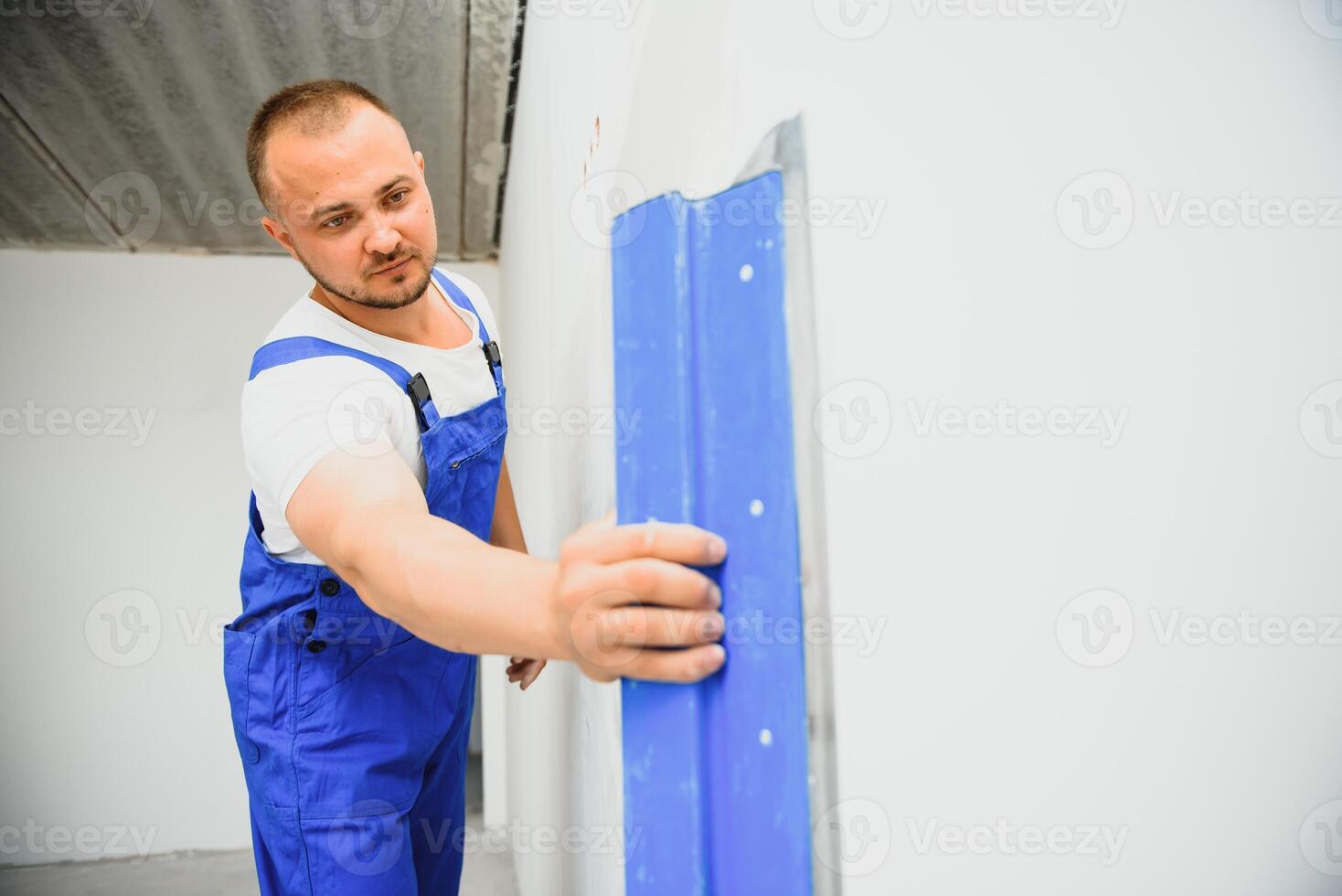 The builder carefully corrects the irregularities of the wall with a trowel. Builder in work clothes against a gray wall. Photo plasterer at work.