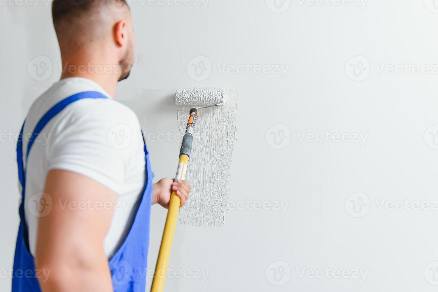Workman in blue overalls painting the wall with a roller, view from the side photo