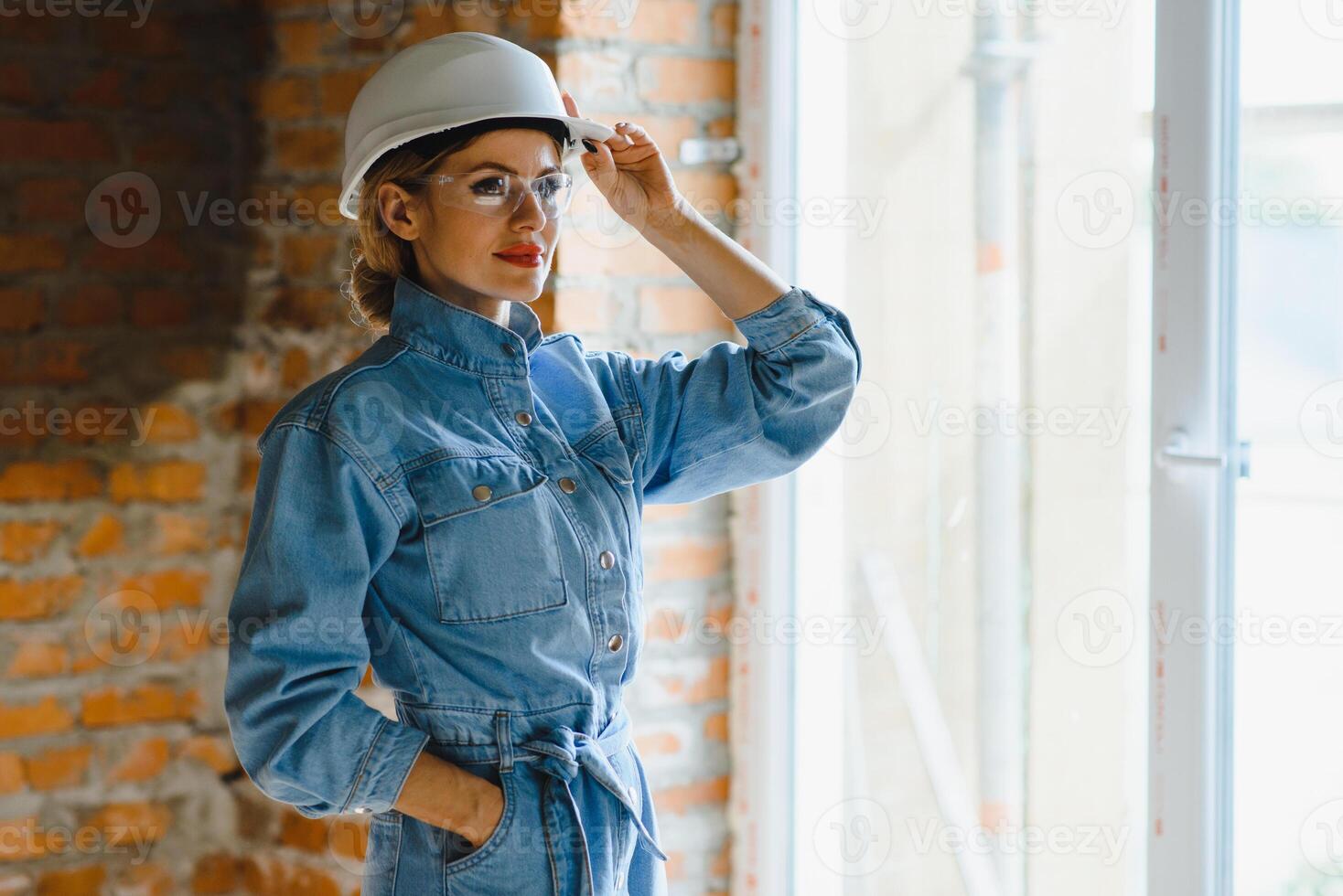 Attractive female construction worker in hardhat. Confident young specialist in checkered blue shirt in jeans standing in empty room. Interior design and renovation service photo