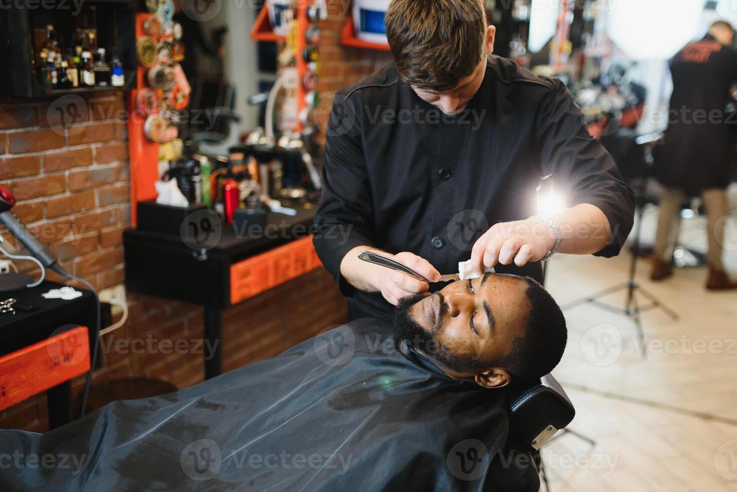 Side view of serious man with stylish modern haircut looking forward in barber shop. Hand of barber keeping straight razor and cutting trendy stripes on head of client. photo