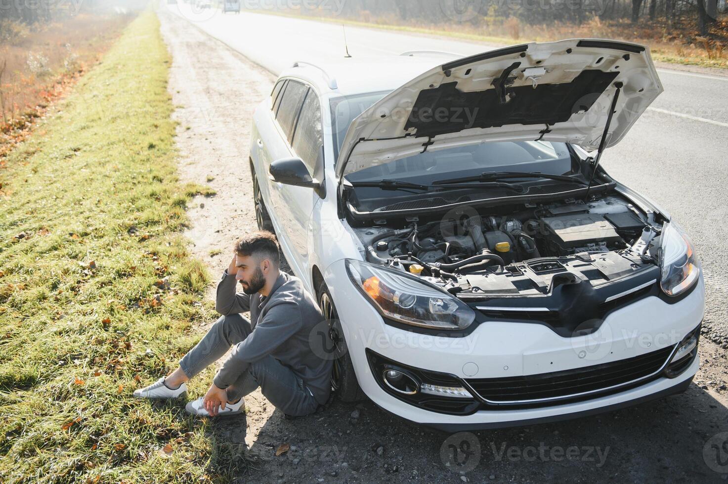 Picture of frustrated man sitting next to broken car with open hood photo