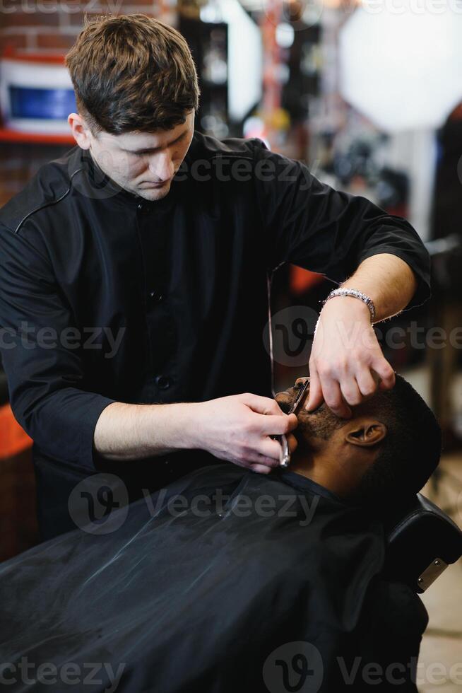 lado ver de grave hombre con elegante moderno Corte de pelo mirando adelante en Barbero tienda. mano de Barbero acuerdo Derecho maquinilla de afeitar y corte de moda rayas en cabeza de cliente. foto