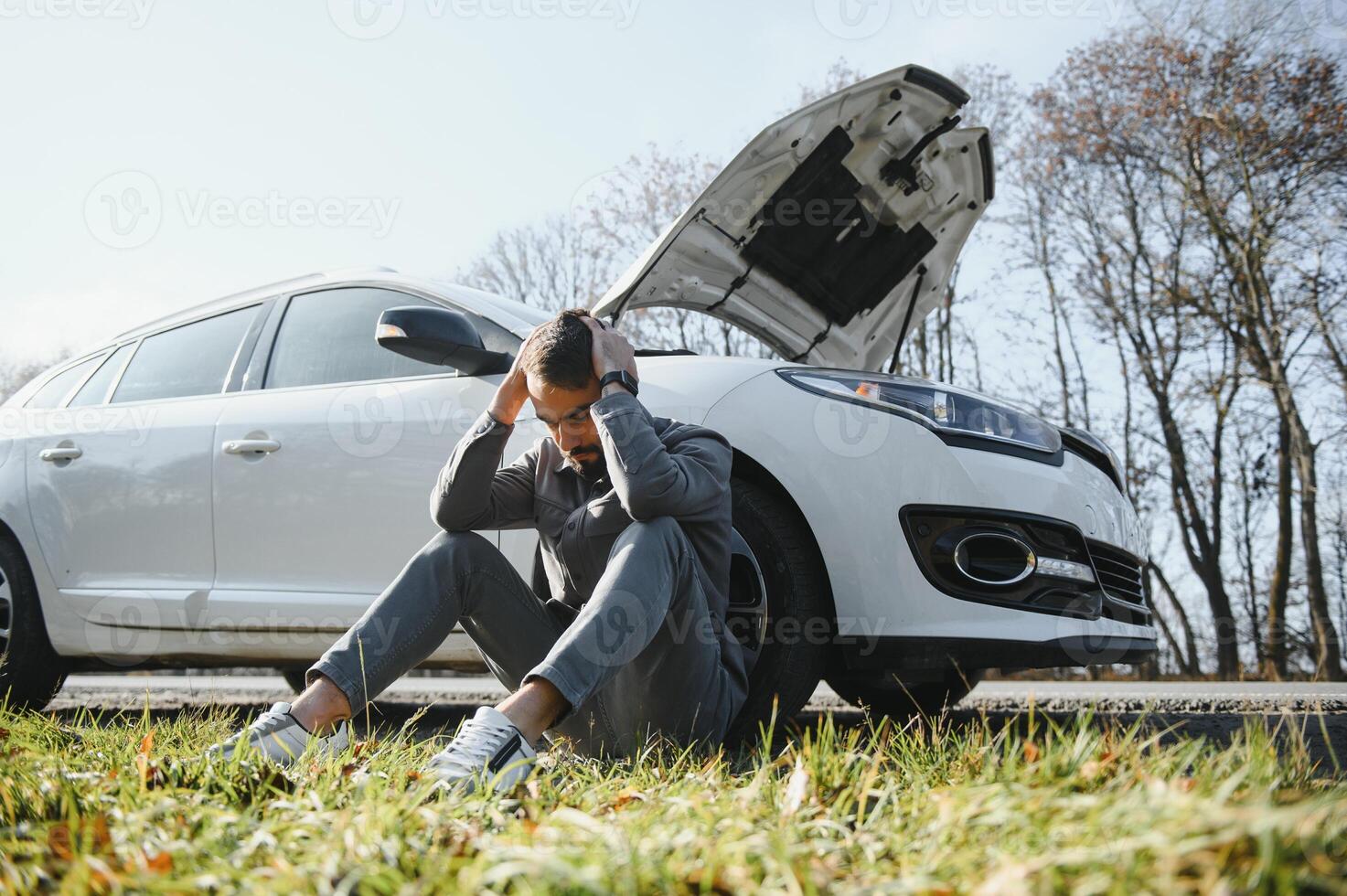 Picture of frustrated man sitting next to broken car with open hood photo