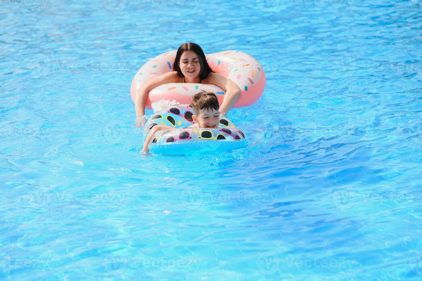 Happy family, young active mother and adorable curly little baby having fun in a swimming pool, child learning to swim in an inflatable toy ring, enjoying summer vacation at a tropical resort photo