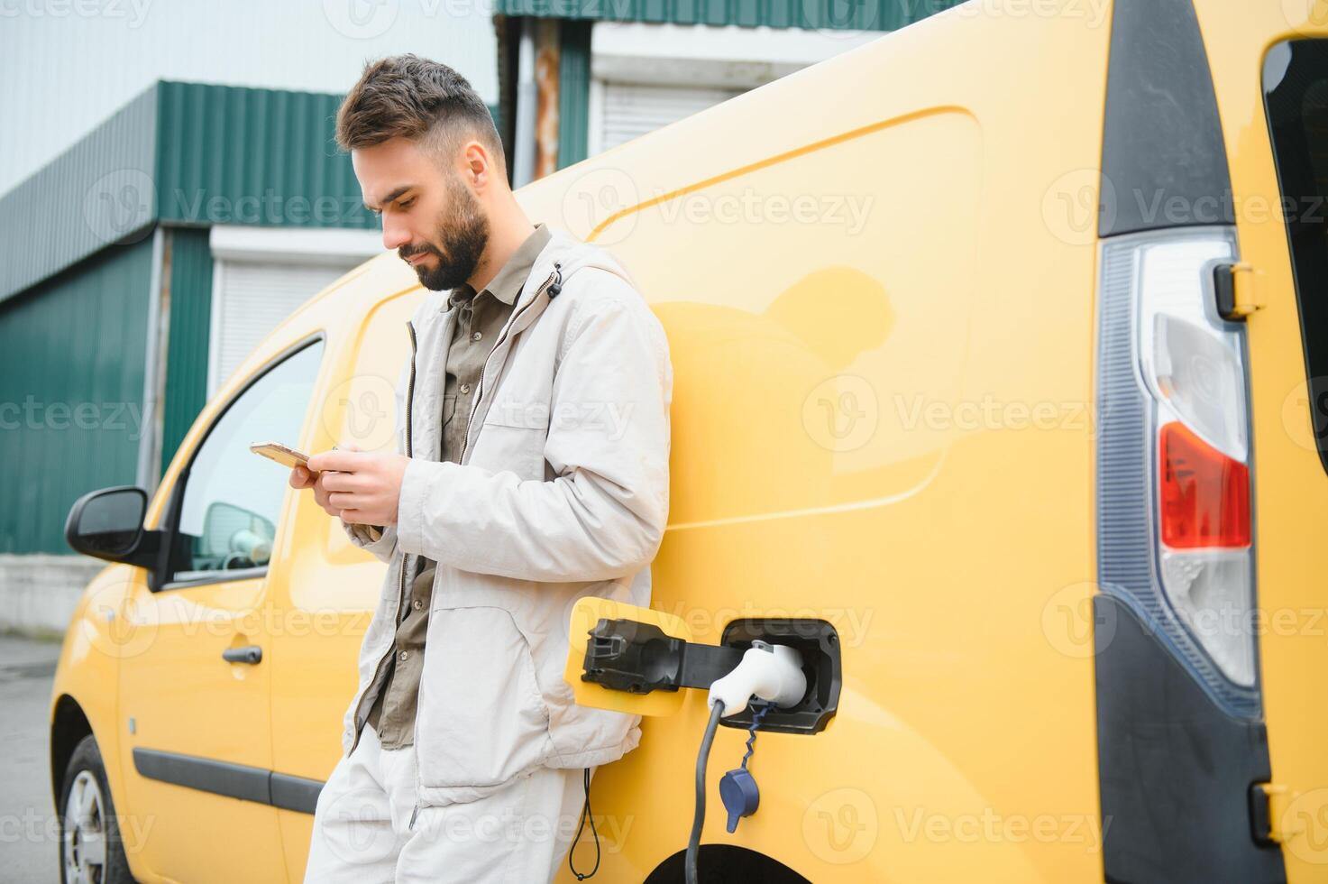 bearded caucasian man standing near an electric car that is charging and making time adjustments on a smartphone. photo