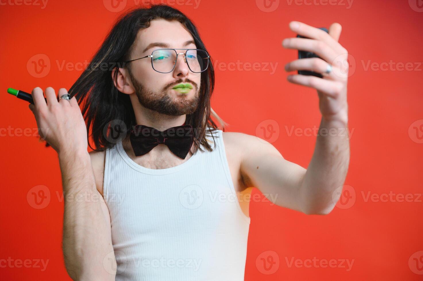 Young man applying lip gloss on his lips and looking in camera like mirror. Gay man doing make up against red background photo