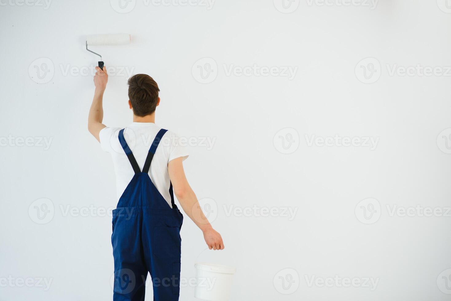 young man in blue overalls painting wall into white color with a roller photo