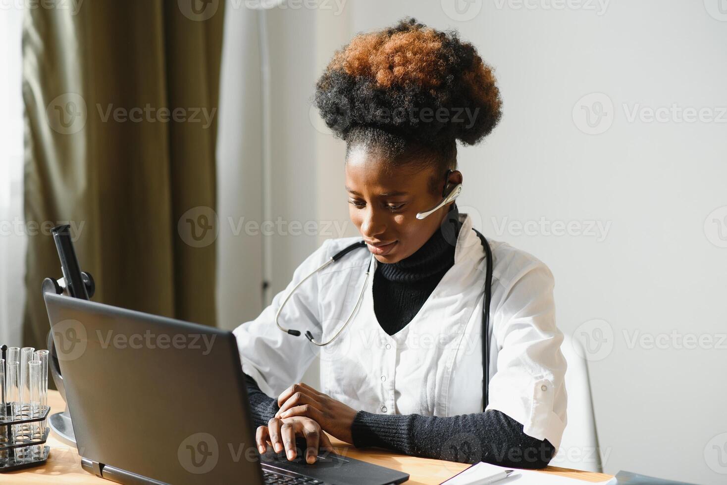 Smiling african american female doctor gp wears white medical coat using laptop computer at workplace gives remote online consultation, working on pc, consulting patient in internet telemedicine chat photo