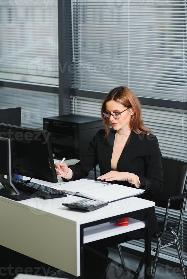 Pretty, nice, cute, perfect woman sitting at her desk on leather chair in work station, wearing glasses, formalwear, having laptop and notebook on the table photo