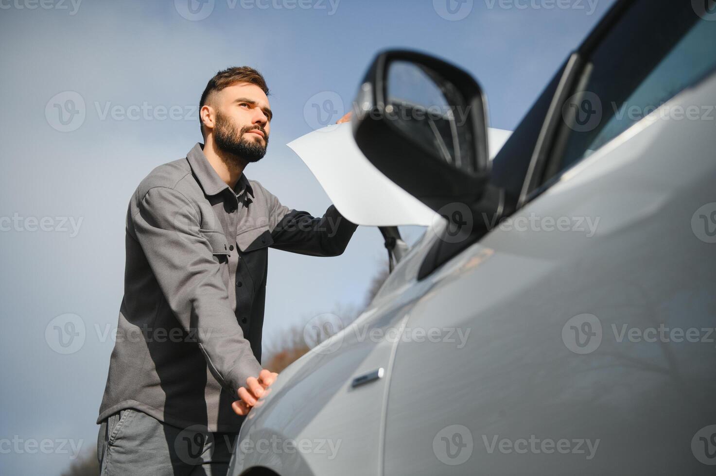 A young man near a broken car with an open hood on the roadside. photo