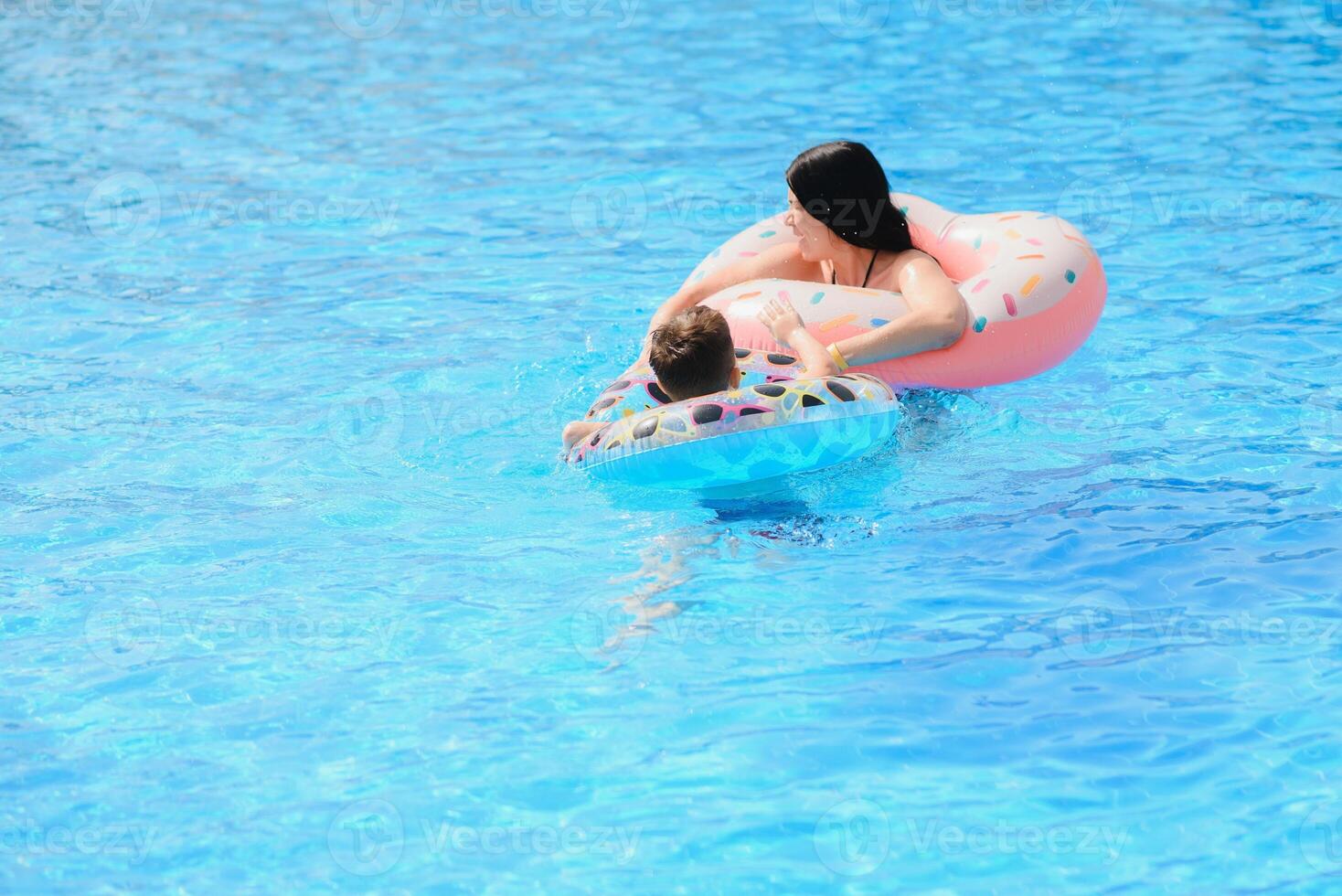 contento familia, joven activo madre y adorable Rizado pequeño bebé teniendo divertido en un nadando piscina, niño aprendizaje a nadar en un inflable juguete anillo, disfrutando verano vacaciones a un tropical recurso foto