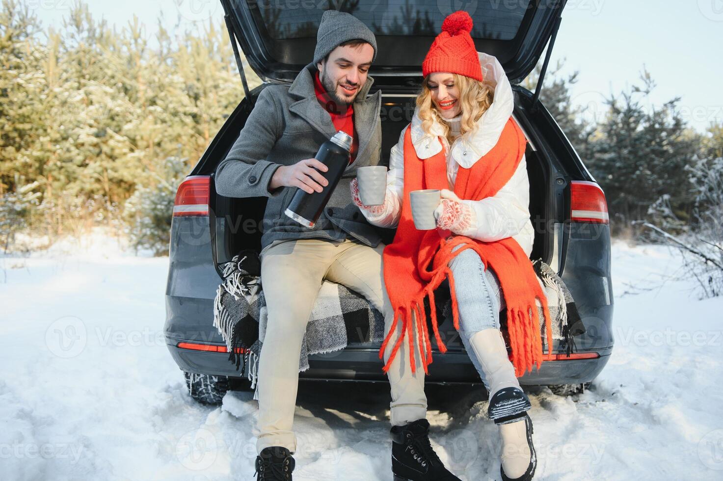 Couple in love sitting in car trunk drinking hot tea in snowy winter forest and chatting. People relaxing outdoors during road trip. Valentines day photo