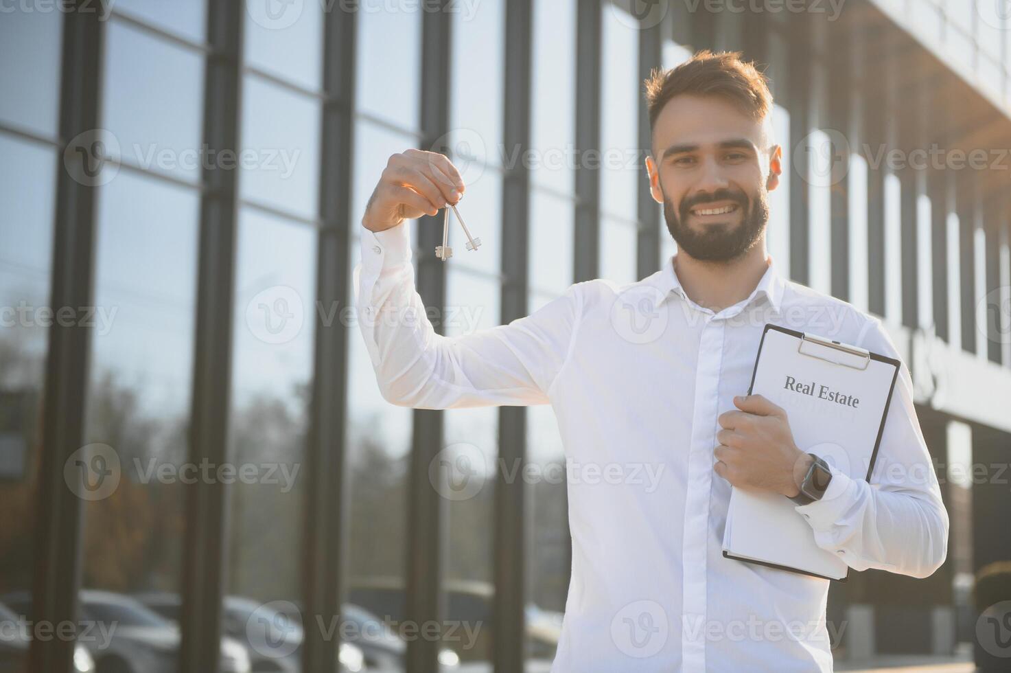 Real estate attractive agent beautiful man with house keys in front of sold apartment, in city street. Young caucasian guy in white shirt with keys just bought new apartment. photo