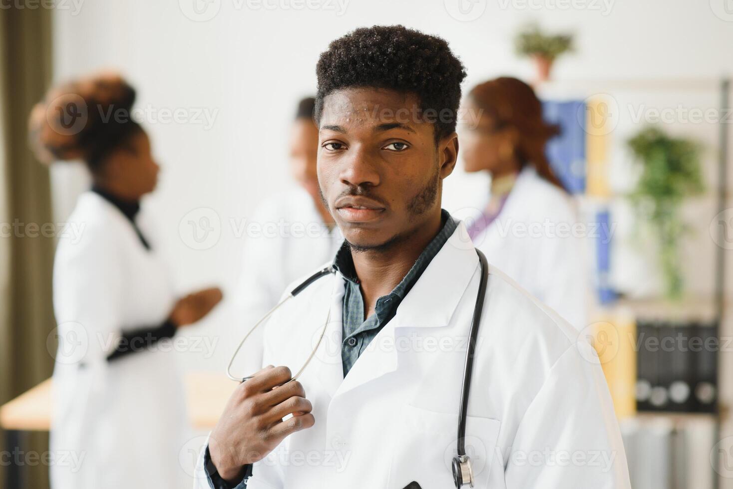 Young African male doctor smiling while standing in a hospital corridor with a diverse group of staff in the background. photo