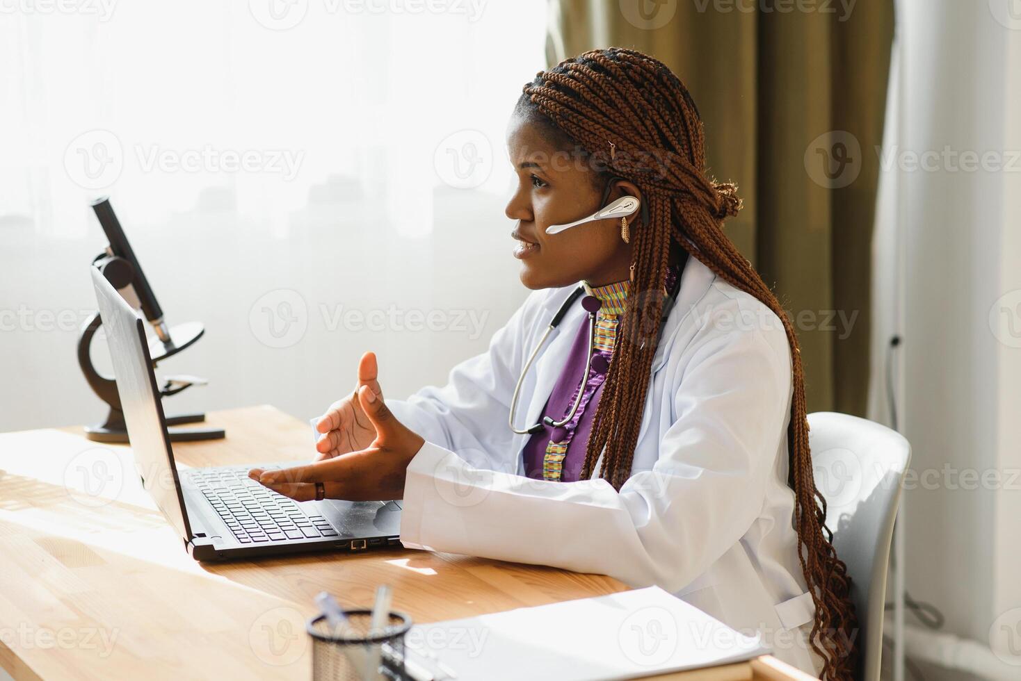 Smiling african american female doctor gp wears white medical coat using laptop computer at workplace gives remote online consultation, working on pc, consulting patient in internet telemedicine chat photo