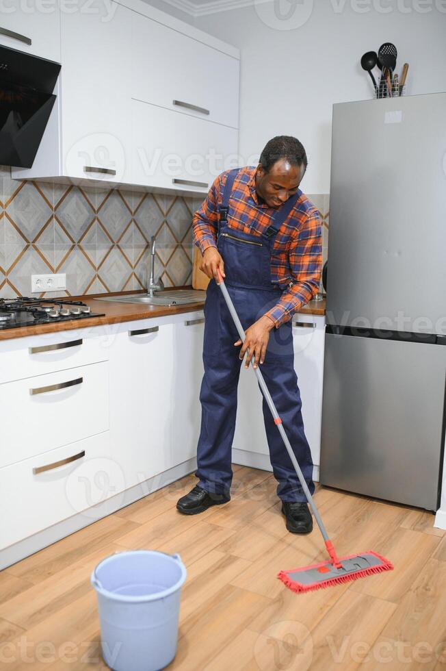 profesional limpiador en azul uniforme Lavado piso y limpiando polvo desde el mueble en el vivo habitación de el departamento. limpieza Servicio concepto foto