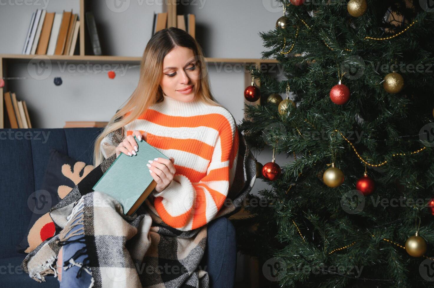 Attractive young lady with dark hair reading interesting book while sitting on grey couch. Blur background of beautiful christmas tree. Cozy atmosphere. photo