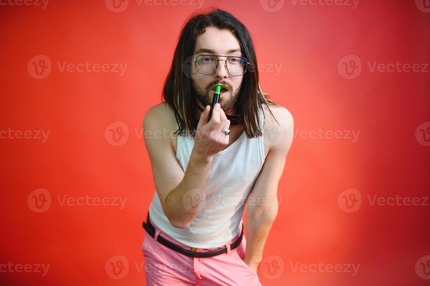Young man applying lip gloss on his lips and looking in camera like mirror. Gay man doing make up against red background photo