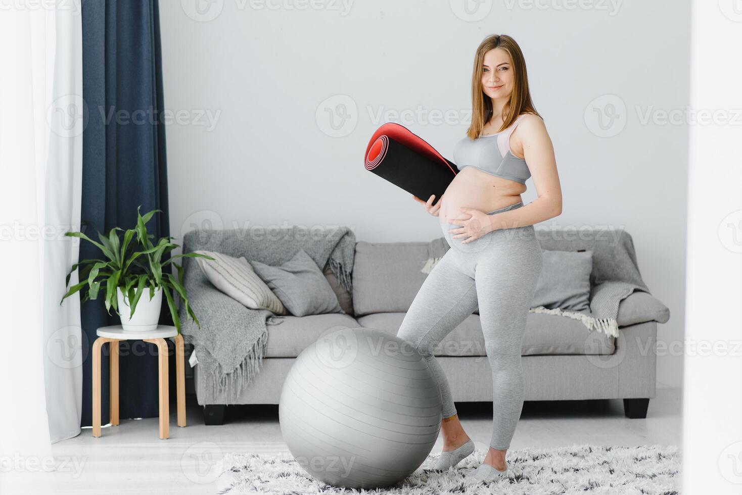 Portrait of a beautiful pregnant woman smiling and holding a rolled yoga mat under her arm. Adorable expectant mother touching her belly and preparing to start her home workout photo