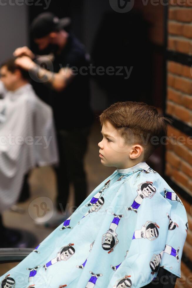 Side view of cute little boy getting haircut by hairdresser at the barbershop. photo