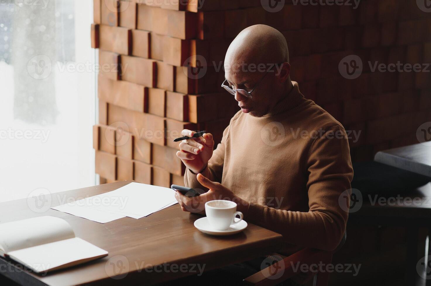 African businessman talking on phone sitting at cafe table, busy entrepreneur working distantly in coffee house with laptop papers speaking on mobile, black man making call having lunch in cafeteria. photo