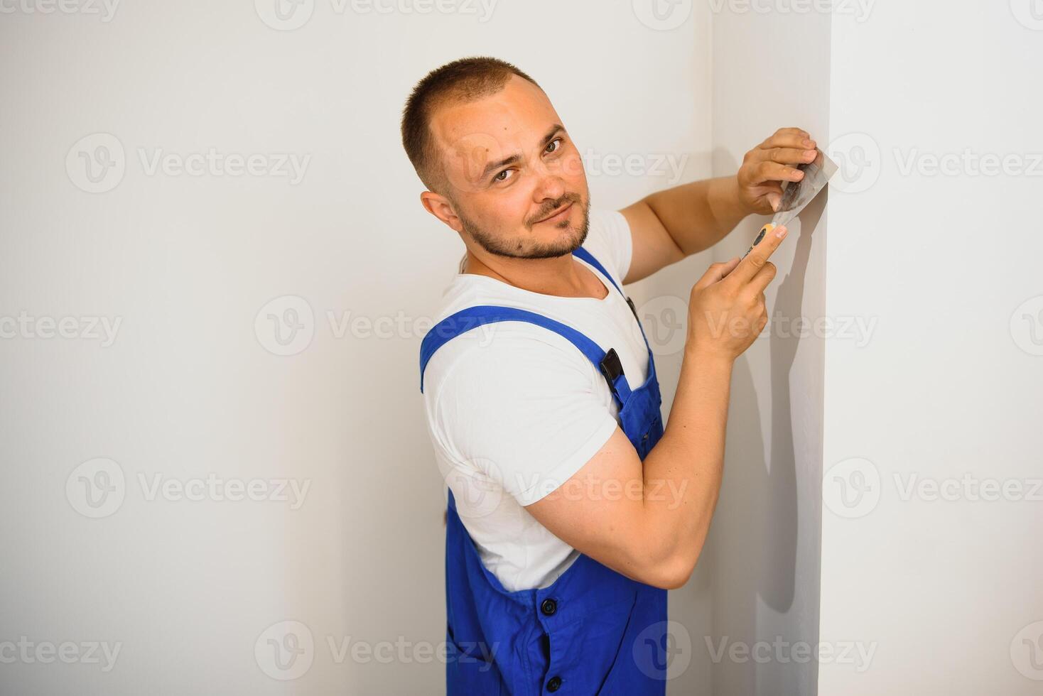 The builder carefully corrects the irregularities of the wall with a trowel. Builder in work clothes against a gray wall. Photo plasterer at work.