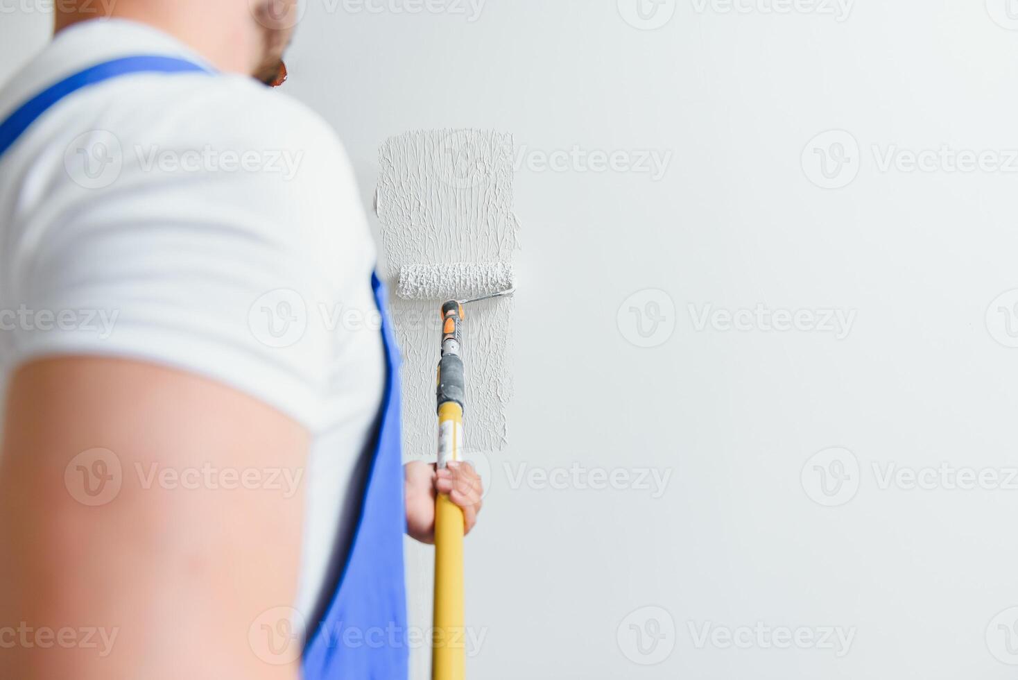 Workman in blue overalls painting the wall with a roller, view from the side photo