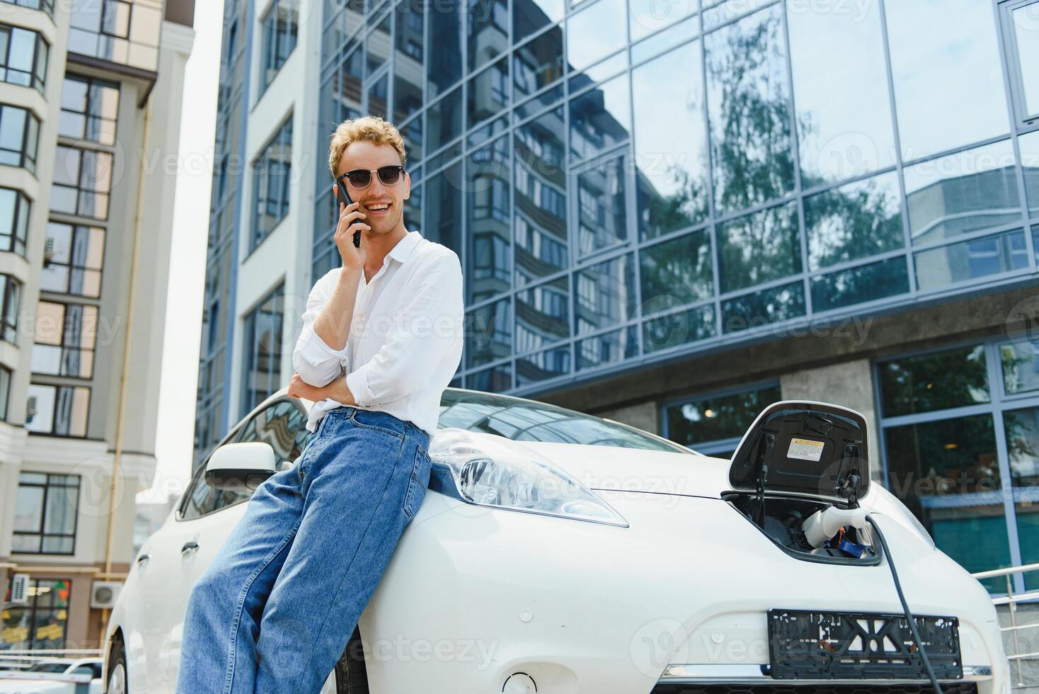 The guy sat down on the hood of the car. His car is charging at the charging station. A man looks at the smartphone screen and smiles. photo