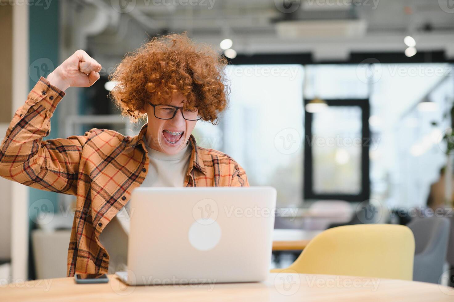 Great news. Male teenager expressing success in front of laptop at cafe, clenching his fist and yelling, copy space. photo