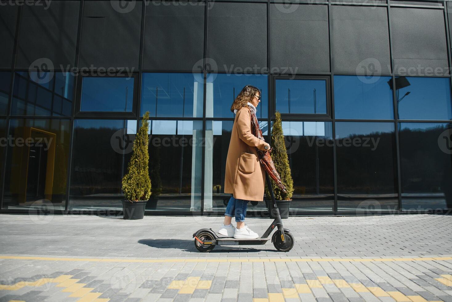 joven hermosa mujer en un chaqueta sonrisas y paseos un eléctrico scooter a trabajo a lo largo oficina edificios foto