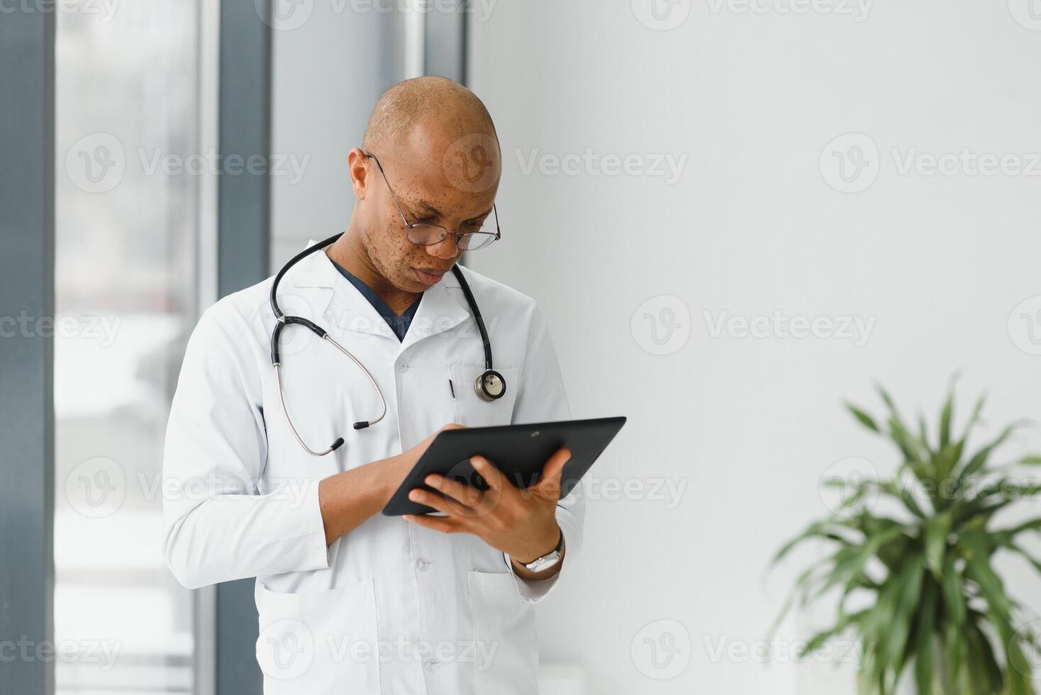 Mature african doctor using digital tablet in corridor . Portrait of confident male doctor using tablet computer in clinic with copy space. Successful smiling doctor in labcoat wearing stethoscope photo