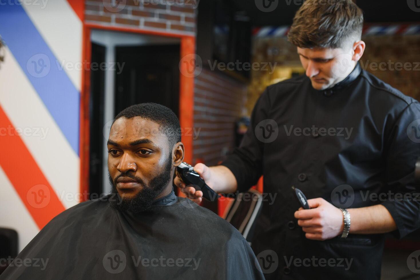 Portrait of young black man being trimmed with professional electric clipper machine in barbershop.Male beauty treatment concept. Young African guy getting new haircut in barber salon photo