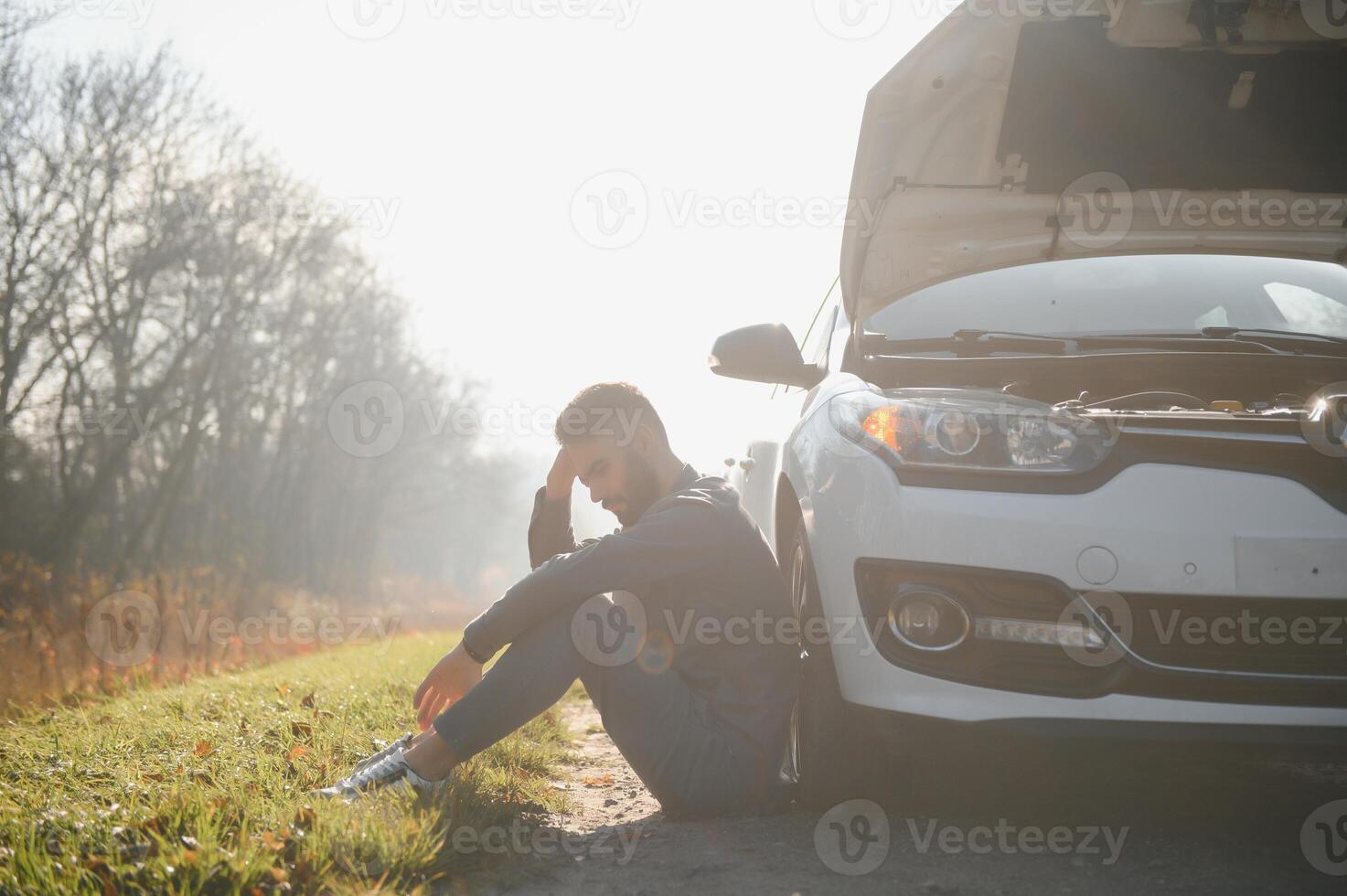 Picture of frustrated man sitting next to broken car with open hood photo