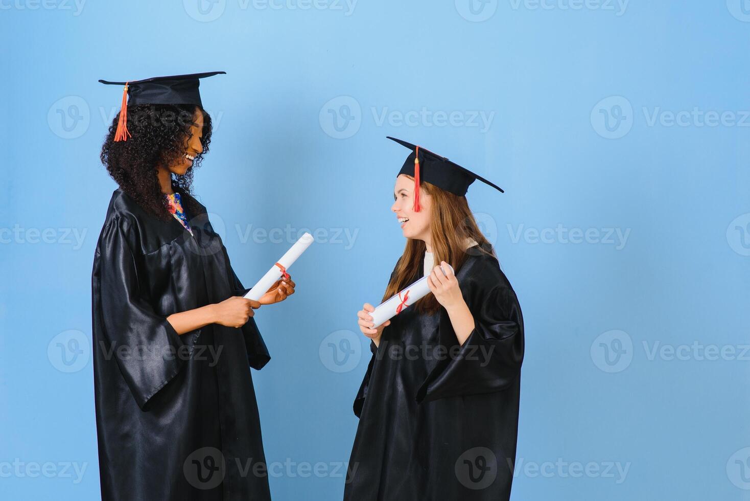 Two girls are posing for take photo in black gowns and hold diploma certificate. They are graduates and hold diploma certificate. They are happy and in good mood.