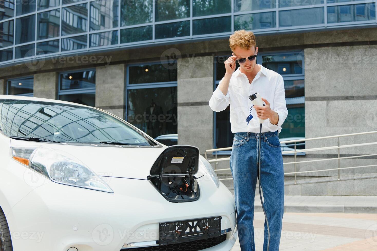 young handsome man holding charging cable at electric charging station point standing near his new car , looking satisfied. photo