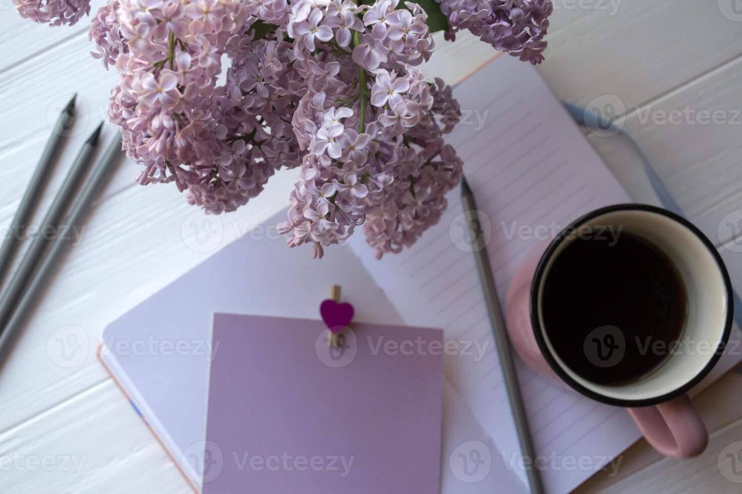 Notepad with pencil, lilac in vase and a cup of coffee on a white wooden desktop. Inspirational workplace. photo