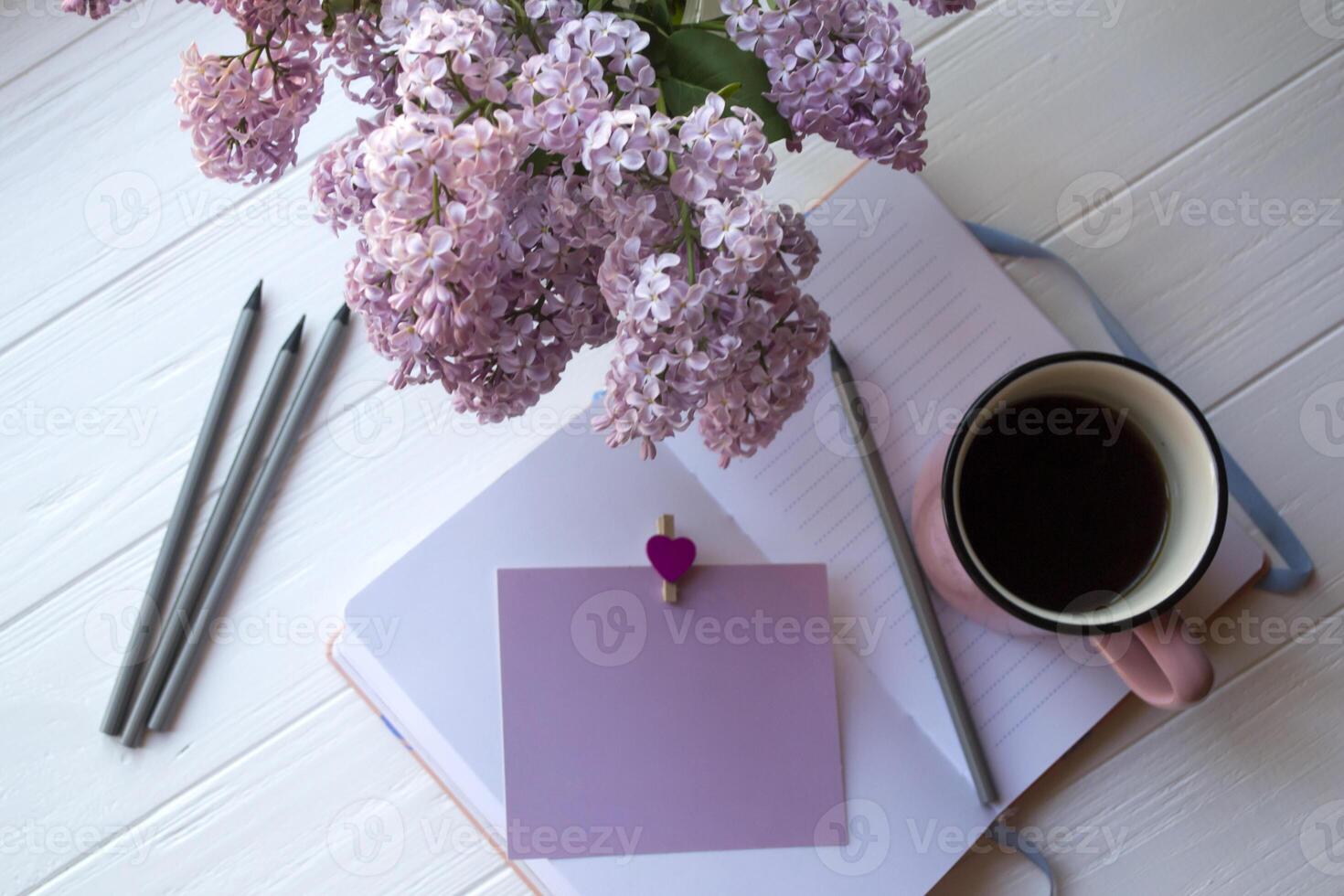 Notepad, pencils, lilac in vase and a cup of coffee on a white wooden desktop. Inspirational workplace. photo