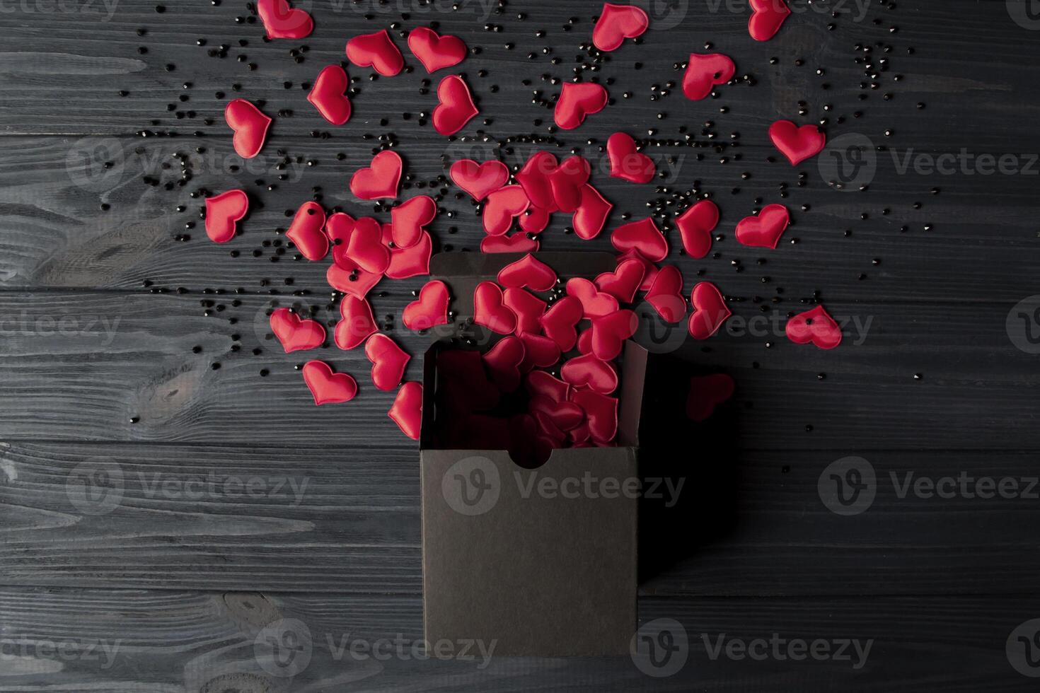 Red hearts are poured out of a black gift box on a dark blue wooden table. photo