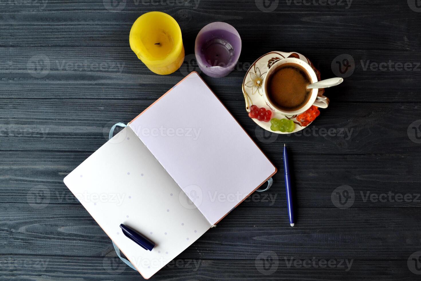 un taza de café con jalea dulces, brillante velas, bolígrafo y abrió cuaderno en el oscuro azul de madera mesa. foto