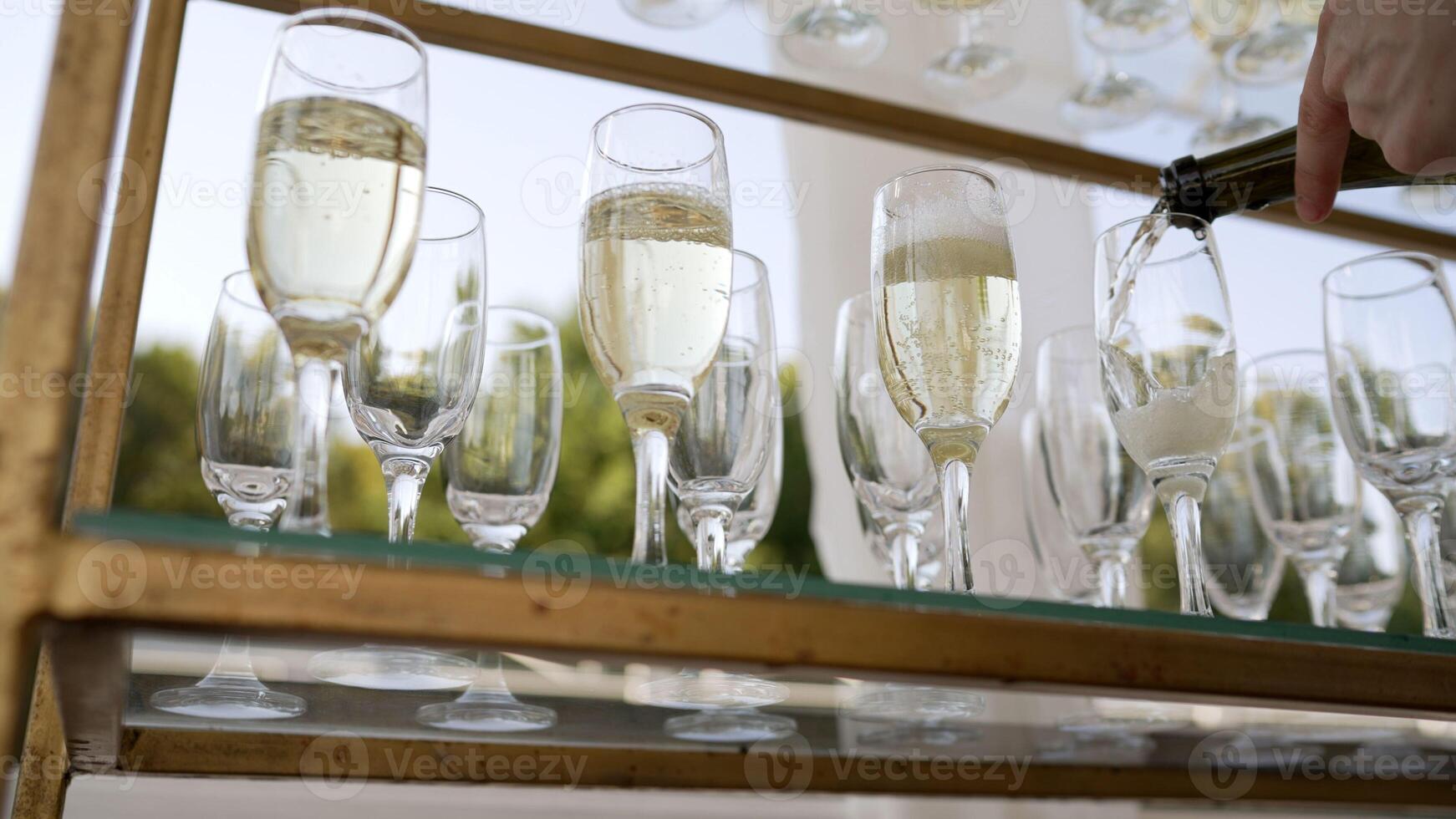 A bottle of sparkling champagne and glasses are poured into a glass at an outdoor wedding event. Open catering, champagne is poured into a glass, close-up. The waiter pours champagne into a glass. photo