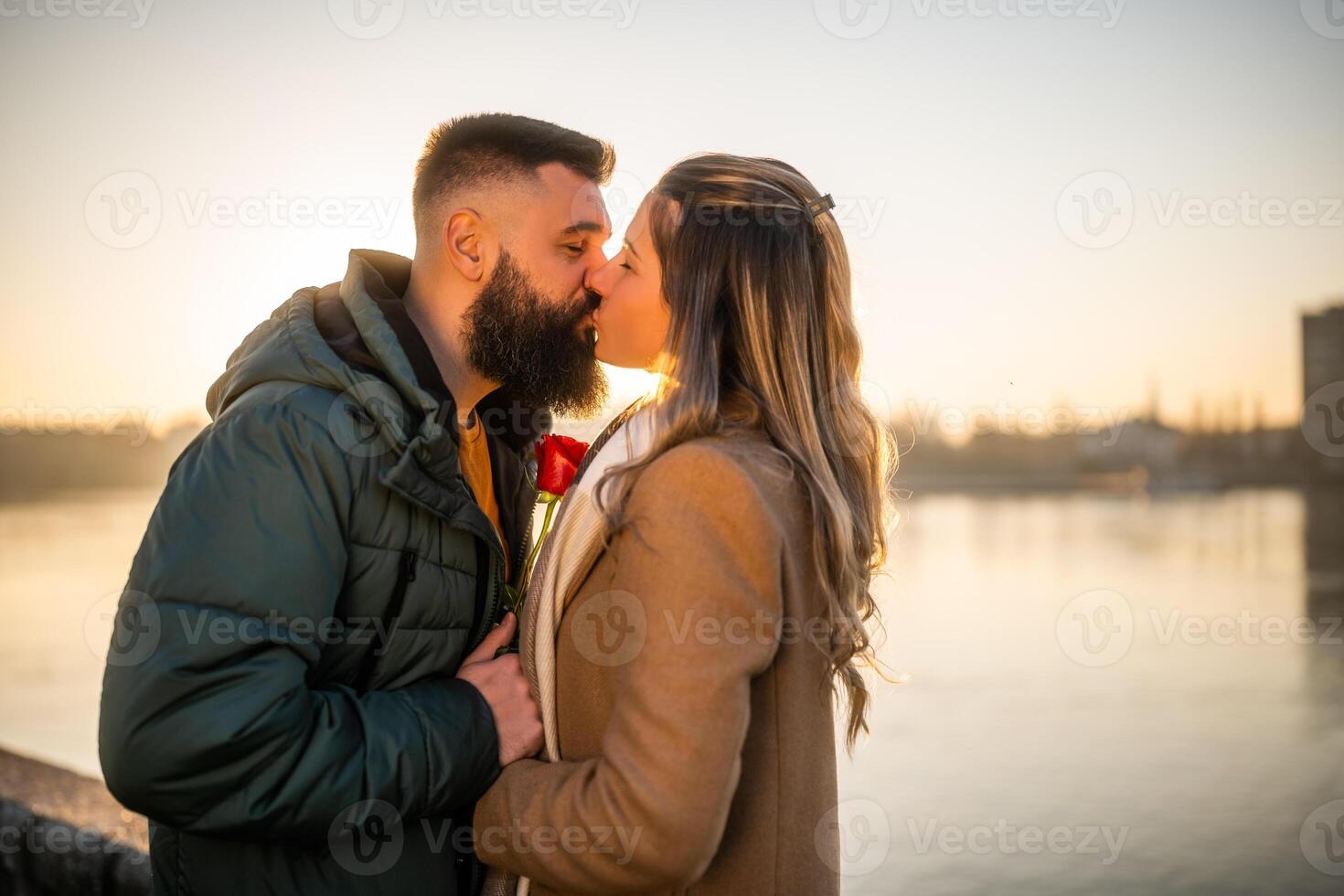 Happy man giving red rose to his woman while they enjoy kissing on a sunset. photo